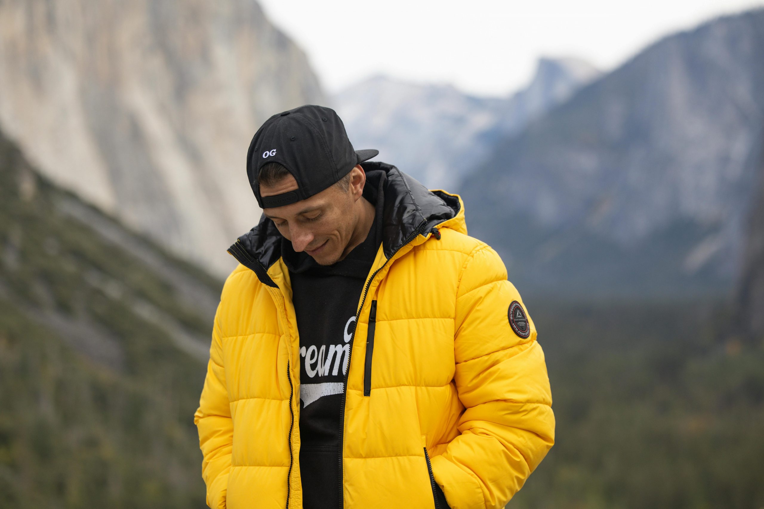 man in a yellow down jacket and cap, standing outdoors with mountains in the distance